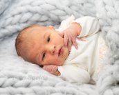 Neugeborenes Baby liegt auf einer weichen, grauen Decke und schaut direkt in die Kamera, mit einer Hand am Gesicht.
