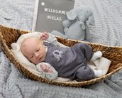 Neugeborenes Baby in einem Korb, trägt einen grauen Overall. Neben ihm steht ein Stoffelefant und ein Schild mit der Aufschrift „Willkommen Niklas“.