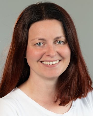 Eine Frau mit langen, braunen Haaren und einem freundlichen Lächeln trägt ein weißes T-Shirt. Sie steht vor einer neutralen grauen Wand und blickt direkt in die Kamera.