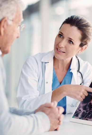 Eine Ärztin zeigt einem älteren Mann auf einem Tablet eine medizinische Bildgebung, während sie auf einen bestimmten Bereich deutet. Der Mann hört aufmerksam zu und hat die Hände gefaltet.