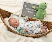 Willkommen Emmett steht auf einem Schild neben einem schlafenden Baby in einem geflochtenen Korb. Das Baby trägt einen bunten Schlafanzug und hat eine entspannte Haltung. Ein grünes Stofftier in Form eines Frosches ist ebenfalls im Bild.
