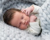 Ein schlafendes Neugeborenes mit dunklem Haar, eingewickelt in eine weiche, graue Decke, liegt auf einem hellen Untergrund.