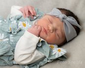 Schlafendes Baby mit einem grauen Haarband und einem blauen Kleid mit Blumenmuster, das auf einem hellen Hintergrund liegt.