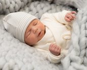 Ein schlafendes Neugeborenes in einem weißen Strickpullover und einer gestreiften Mütze, eingekuschelt in eine graue Decke.