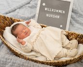 Neugeborenes Baby in einem Korb, gekleidet in einem weißen Kleid, mit einem Schild, das "Willkommen Nursan" zeigt.