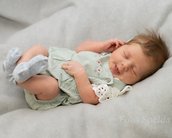 Ein schlafendes Baby in einem grünen Overall mit weißen Details und grauen Socken, auf einem weichen, grauen Untergrund.
