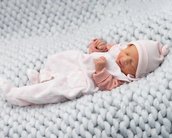 Ein schlafendes Baby in einem rosa-weißen Strampler und einer Mütze liegt auf einer gestrickten Decke in neutralen Farben.
