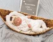 Neugeborenes Baby in einem geflochtenen Korb, trägt einen weißen Schlafanzug mit Punkten und einem Haarband. Ein Schild im Hintergrund begrüßt "Lena Zofia".