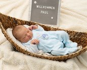 Ein neugeborenes Baby liegt in einem Korb auf einer weichen Decke, trägt einen blauen Schlafanzug mit einem Tiermotiv und hat die Hände an das Gesicht gelegt. Neben dem Baby steht ein Schild mit der Aufschrift "Willkommen Paul".
