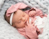 Neugeborenes Baby mit einem rosa Haarband und einem rosa Oberteil, das in einer weichen Decke schläft und an den Fingern saugt.
