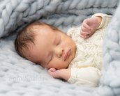 Schlafendes Baby in einem weichen, grauen Strickbett, eingekuschelt in eine Decke, mit einem zarten, gemusterten Oberteil.