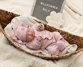 Neugeborenes Mädchen in rosa Kleidung schläft in einem Korb, umgeben von einer Decke. Ein Schild mit "Willkommen Nela" und "Hello World" ist sichtbar.