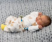 Neugeborenes Baby liegt auf einer grauen, gestrickten Decke, trägt einen weißen Overall mit Tiermotiven und einen bunten Schnullerhalter.