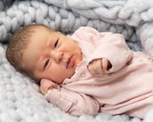 Ein Neugeborenes in einem rosa Strampler liegt auf einer weichen, grauen Decke. Das Baby hat dunkles Haar und schaut direkt in die Kamera, während es eine Hand an die Brust legt.