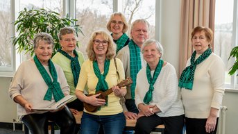 Eine Gruppe von sieben Personen steht um einen Tisch, eine Frau hält eine Ukulele. Alle tragen grüne Schals und lächeln.