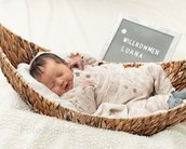 Neugeborenes Baby schläft in einem Korb, trägt einen gestrickten Anzug. Ein Schild mit der Aufschrift "Willkommen Luana" ist sichtbar.