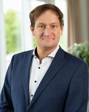 Ein Mann in einem dunkelblauen Anzug mit einem weißen Hemd steht vor einem Fenster, das Tageslicht hereinlässt. Er hat kurzes, braunes Haar und schaut direkt in die Kamera.