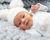 Neugeborenes Baby mit einem weißen Mütze mit Pompons, das auf einem grauen, strukturierten Hintergrund liegt und in die Kamera schaut.