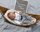 Neugeborenes Baby in einem Korb, trägt gestreifte Kleidung. Ein Schild im Hintergrund zeigt "Willkommen Aron Oskar".