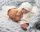 Neugeborenes Baby schläft friedlich in einem weichen, grauen Strickbett, trägt einen weißen Anzug mit Punkten und eine große Schleife.