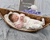 Ein schlafendes Baby liegt in einem geflochtenen Korb, umgeben von einer Decke, mit einem Schild, das "Willkommen Malin" zeigt.