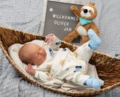 Neugeborenes Baby in einem Korb, trägt einen bedruckten Schlafanzug. Neben ihm steht ein Plakat mit "Willkommen Oliver Jan" und einem Stofftier.