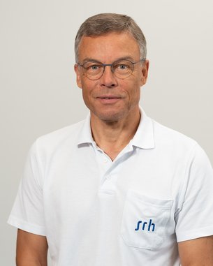 Ein Mann mit Brille trägt ein weißes Polohemd mit dem Logo "srh" auf der Brust. Er steht vor einem neutralen Hintergrund und schaut direkt in die Kamera.