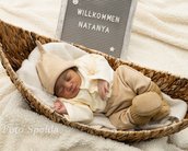 Ein schlafendes Baby in einem Korb, gekleidet in beigem Outfit mit Mütze, neben einem Schild mit der Aufschrift "Willkommen Natanya".