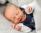 Neugeborenes Baby in blauer Latzhose schläft auf einem grauen Hintergrund und hat die Hände leicht angehoben.