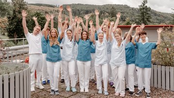 Karriere in Friedrichroda Eine Gruppe von 15 Personen in medizinischer Kleidung hebt die Hände in die Luft, lächelnd, vor einer natürlichen Kulisse mit Bäumen und Hügeln.