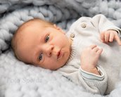 Neugeborenes Baby mit blauen Augen, das auf einer weichen, grauen Decke liegt und in die Kamera schaut. Es trägt einen hellen Pullover.
