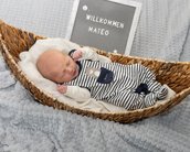 Neugeborenes Baby in einem geflochtenen Korb, trägt einen gestreiften Overall. Ein Schild mit der Aufschrift "Willkommen Mateo" ist im Hintergrund.
