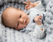 Ein Baby liegt auf einer grauen Decke, trägt einen grauen Strampler und zeigt mit beiden Händen ein Zeichen.