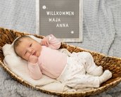 Neugeborenes Baby liegt in einem Korb, trägt einen rosa Body und eine weiße Hose mit Punkten. Neben dem Baby steht ein Schild mit "Willkommen Maja Anna".