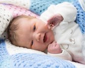 Neugeborenes Baby mit braunen Haaren, das auf einer bunten Decke liegt und lächelt, während es mit einer Hand spielt.