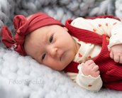 Ein Baby mit einem roten Haarband und einem roten Overall mit Herzen liegt auf einer grauen Decke.