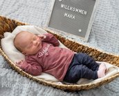 Neugeborenes Baby schläft in einem Korb, trägt ein rosa Oberteil und eine blaue Hose. Ein Schild im Hintergrund begrüßt "Mara".