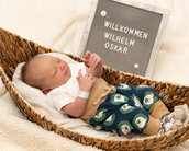 Neugeborenes Baby in einer Wanne, trägt einen weißen Body und eine Hose mit Tiermotiven. Neben ihm steht ein Schild mit dem Text „Willkommen Wilhelm Oskar“.