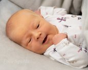 Ein neugeborenes Baby mit einem sanften Lächeln, das in einem weißen Schlafanzug mit bunten Mustern auf einer grauen Decke liegt.