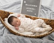 Neugeborenes Baby in einem Korb, trägt einen weißen Schlafanzug mit roten Herzen. Ein Schild im Hintergrund begrüßt "Jasmina-Maria".