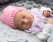 Schlafendes Baby mit rosa Mütze und lila Oberteil, auf einer grauen, gestrickten Decke.
