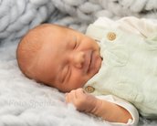Ein schlafendes Baby mit einem leichten Lächeln, gekleidet in einen hellgrünen Overall, liegt auf einer weichen, grauen Decke.