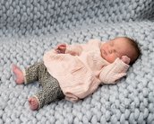 Ein schlafendes Baby in einem rosa Pullover und gepunkteten Leggings liegt auf einer grauen, gestrickten Decke.