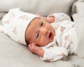 Neugeborenes Baby mit einem weißen Hut und einem gestreiften Bodysuit, das auf einer grauen Decke liegt und die Hände an den Kopf legt.
