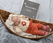 Neugeborenes Mädchen in einem Korb, trägt ein blumiges Outfit und eine Schleife, neben einem Schild mit der Aufschrift "Willkommen Ella".