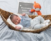 Neugeborenes Baby in einem Korb, trägt eine hellblaue Weste und gestreifte Hose. Neben ihm steht ein Schild mit "Willkommen Mario Alexandru" und ein Plüschtier.