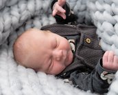 Ein schlafendes Baby liegt in einer weichen, grauen Decke, trägt einen dunkelgrauen Pullover mit einem Muster und hat die Hände leicht angehoben.