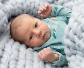 Neugeborenes Baby mit blauen Augen und hellem Haar, eingekuschelt in eine graue, dicke Decke, trägt einen grünen Bodysuit mit bunten Motiven.