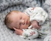 Ein schlafendes Neugeborenes liegt auf einer weichen, grauen Decke. Es trägt einen weißen Schlafanzug mit Dinosaurier- und Pflanzenmotiven. Das Baby hat leicht gewellte Haare und eine entspannte Körperhaltung.