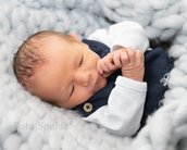 Neugeborenes Baby mit braunen Haaren, das in einer grauen Decke liegt und seine Hände zusammenhält. Es trägt einen blauen Overall mit weißen Ärmeln.
